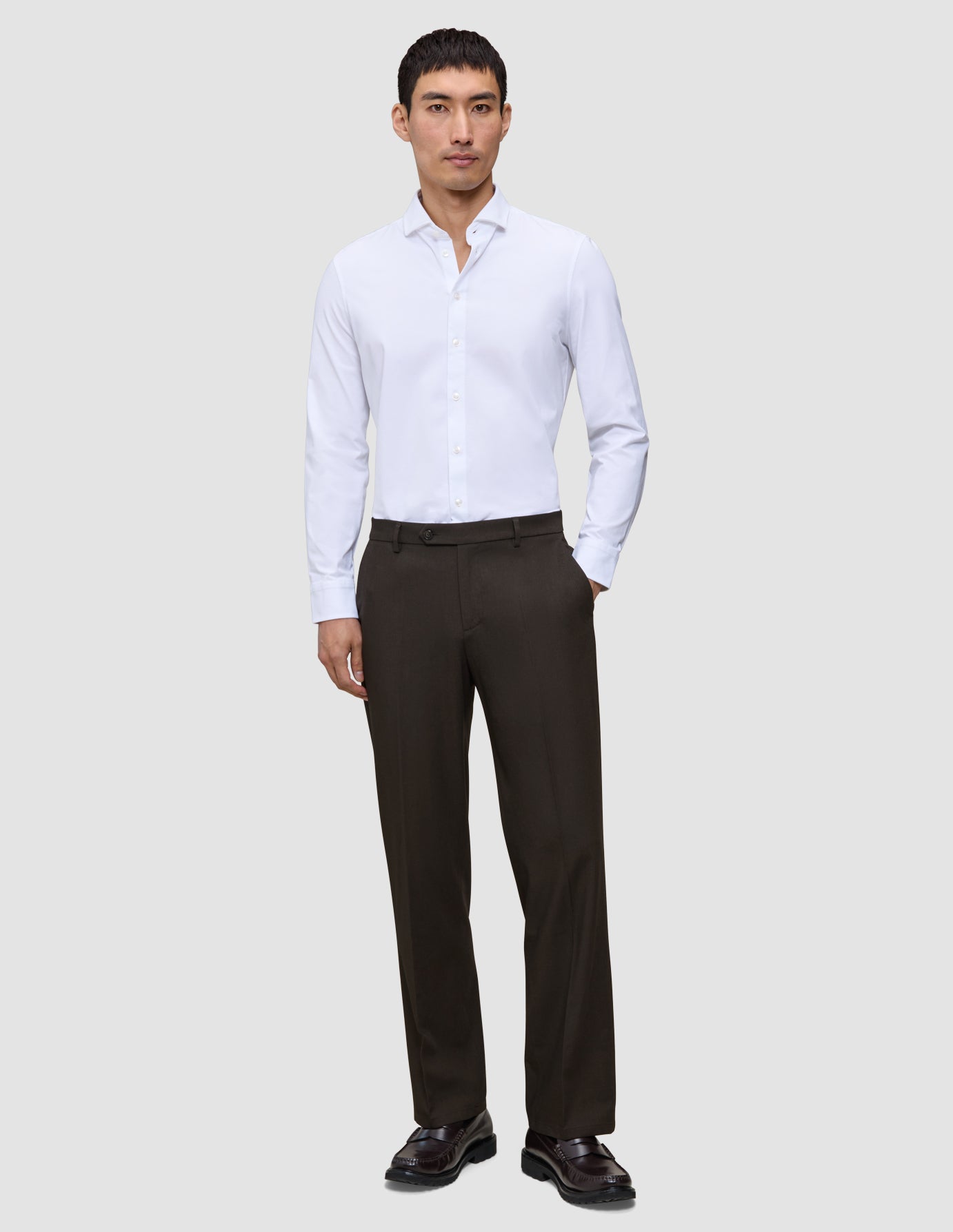 Ein Mann, der ein weißes, langärmeliges Hemd mit Knöpfen und eine Motion Suit Pants Relaxed Cognac sowie dunkle Loafer trägt.